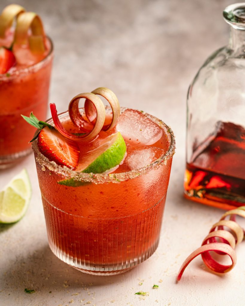 a rhubarb - strawberry cocktail with a curly rhubarb, strawberry and lime wedge garnish