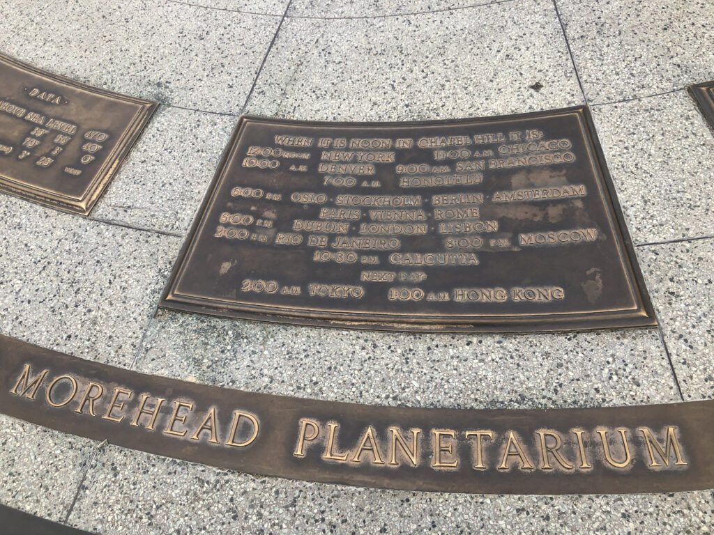 Inscription on the sundial at the Morehead Planetarium reading “When it is noon in Chapel Hill, it is 12:00 noon New York, 11:00am Chicago, 10:00am Denver, 9:00am San Francisco, 7:00am Honolulu, 6:00pm Oslo, Stockholm, Berlin, Amsterdam, Paris, Vienna, Rome, 5:00pm Dublin, London, Lisbon, 2:00pm Rio de Janeiro, 8:00pm Moscow, 10:30pm Calcutta, next day 2:00am Tokyo, 1:00am Hong Kong”